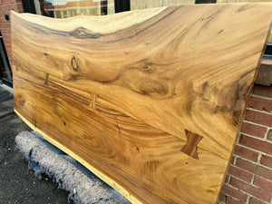 A close-up of a live edge acacia wood slab dining table top, showcasing its natural grain patterns and rich brown tones with unique, organic edges. The wood surface is smooth and polished, highlighting the wood's rustic yet refined beauty.