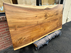 A close-up of a live edge acacia wood slab dining table top, showcasing its natural grain patterns and rich brown tones with unique, organic edges. The wood surface is smooth and polished, highlighting the wood's rustic yet refined beauty.