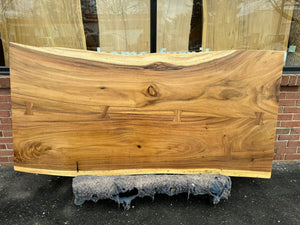 A close-up of a live edge acacia wood slab dining table top, showcasing its natural grain patterns and rich brown tones with unique, organic edges. The wood surface is smooth and polished, highlighting the wood's rustic yet refined beauty.