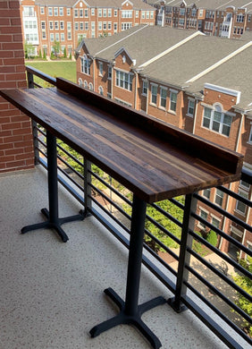 A teak outdoor bar table with a black metal frame, positioned on a balcony with an apartment building in the background.